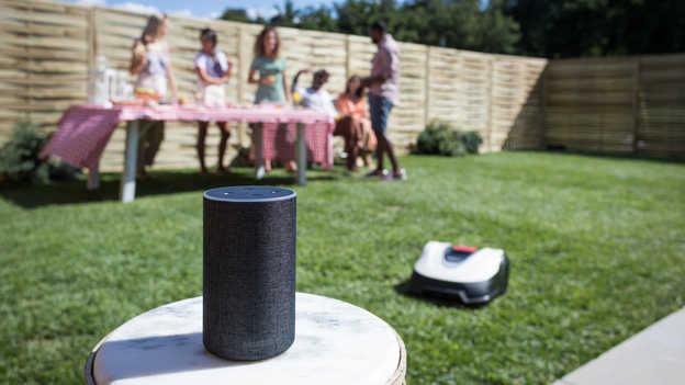 Detailný pohľad na zariadenie Amazon Alexa s kosačkou Miimo a záhradnou párty na trávniku v pozadí.