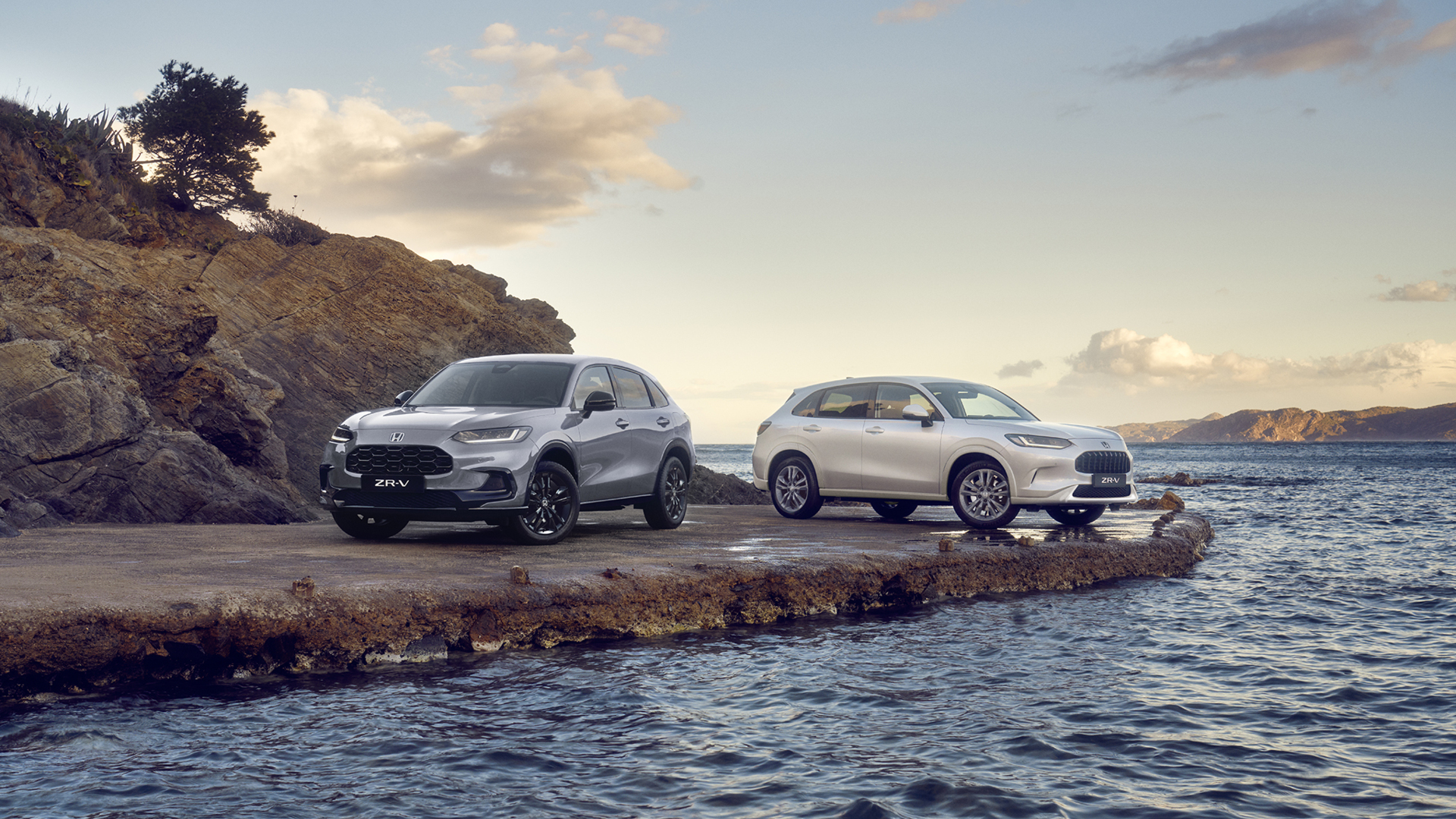 Two Honda ZR-V SUVs, one blue and one white, parked on a rocky coastal outcrop with water and hills in the background under a partly cloudy sky.
