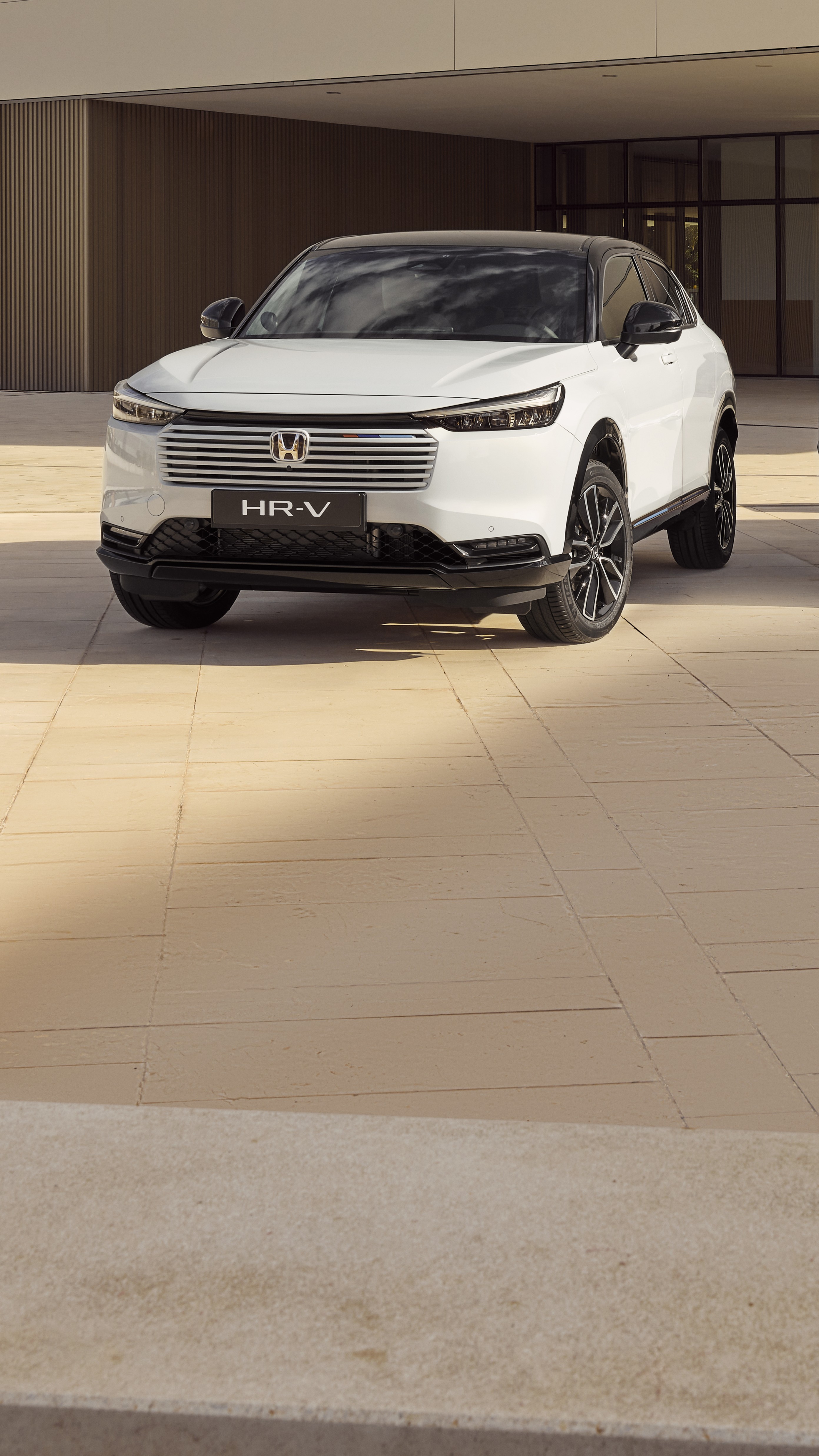 Biele SUV Honda HR-V zaparkované pred modernou budovou s béžovými obkladanými stenami. Vozidlo je umiestnené pod uhlom, čo predvádza jeho elegantný dizajn a osobititú prednú mriežku.