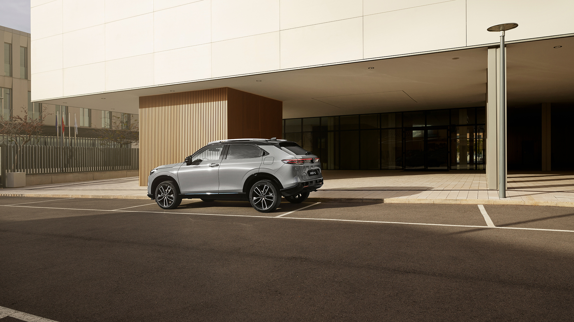Strieborná Honda HR-V je zaparkovaná na vonkajšom parkovacom mieste pred modernou budovou so sklenenými stenami a zvislým dreveným obložením, fotografovaná zo zadného trojštvrťového uhla za denného svetla.