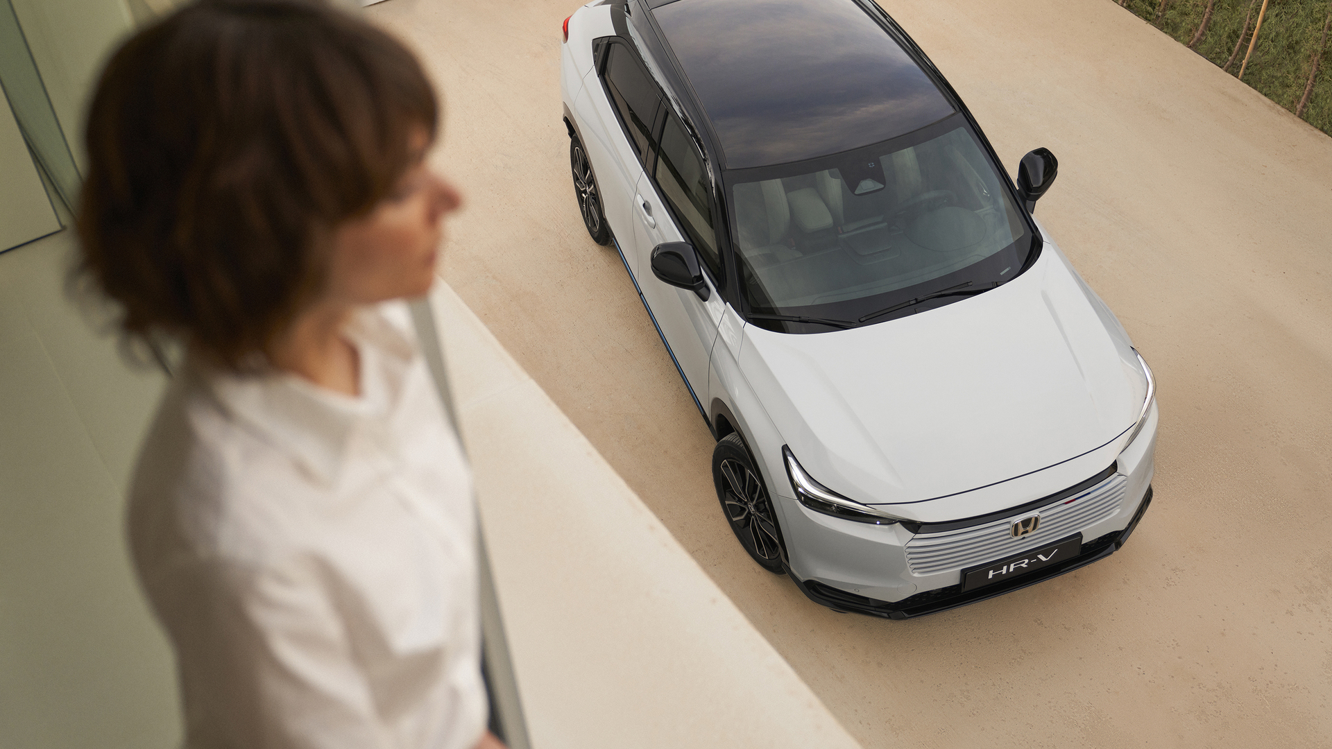 Pohľad zhora na biely Honda HR-V s čiernou strechou zaparkovaný na svetlej príjazdovej ceste, zatiaľ čo rozmazaná osoba stojí v interiéri v popredí a pozerá sa smerom k autu.