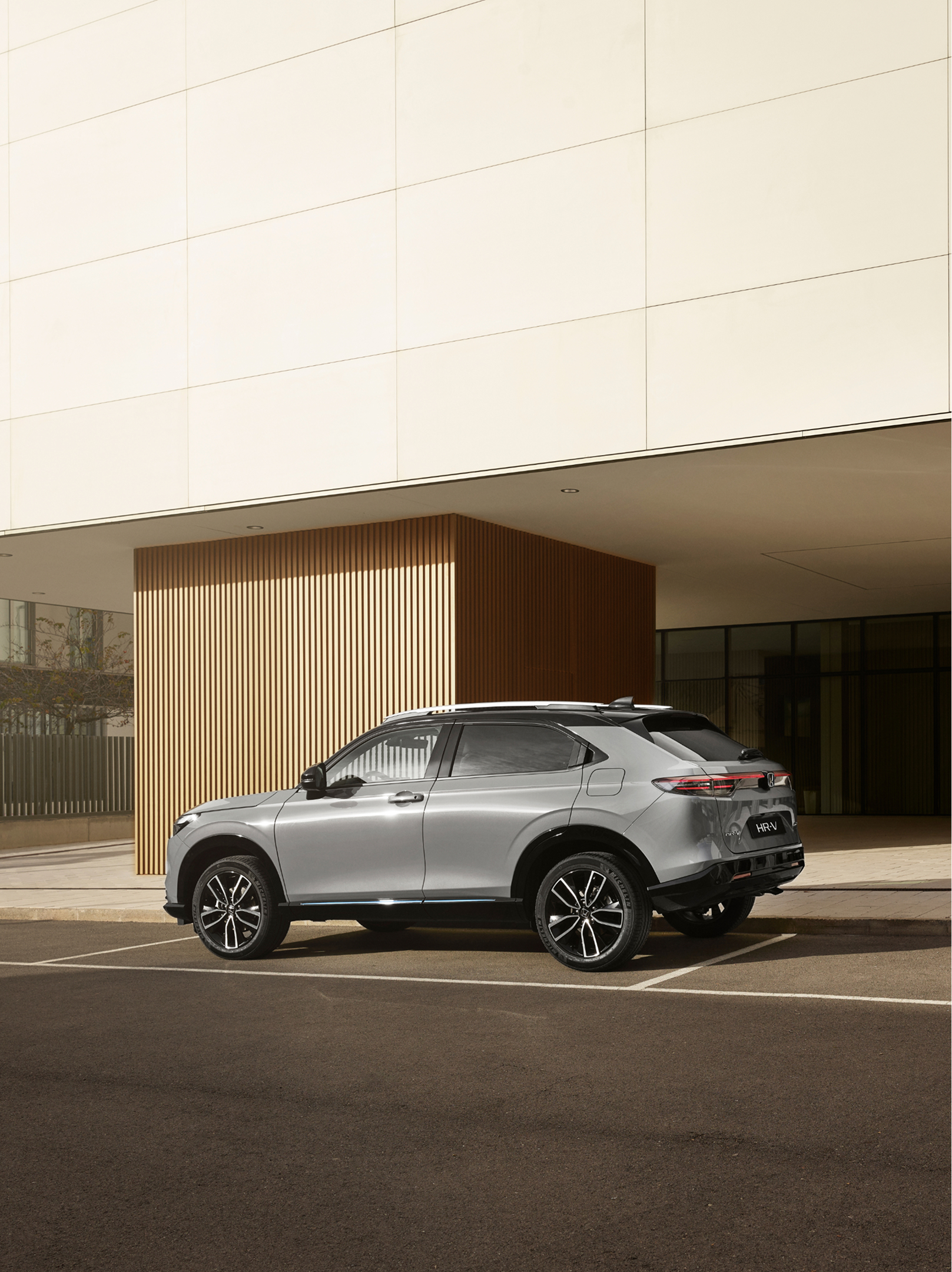 Strieborná Honda HR-V je zaparkovaná na vonkajšom parkovacom mieste pred modernou budovou so sklenenými stenami a zvislým dreveným obložením, fotografovaná zo zadného trojštvrťového uhla za denného svetla.