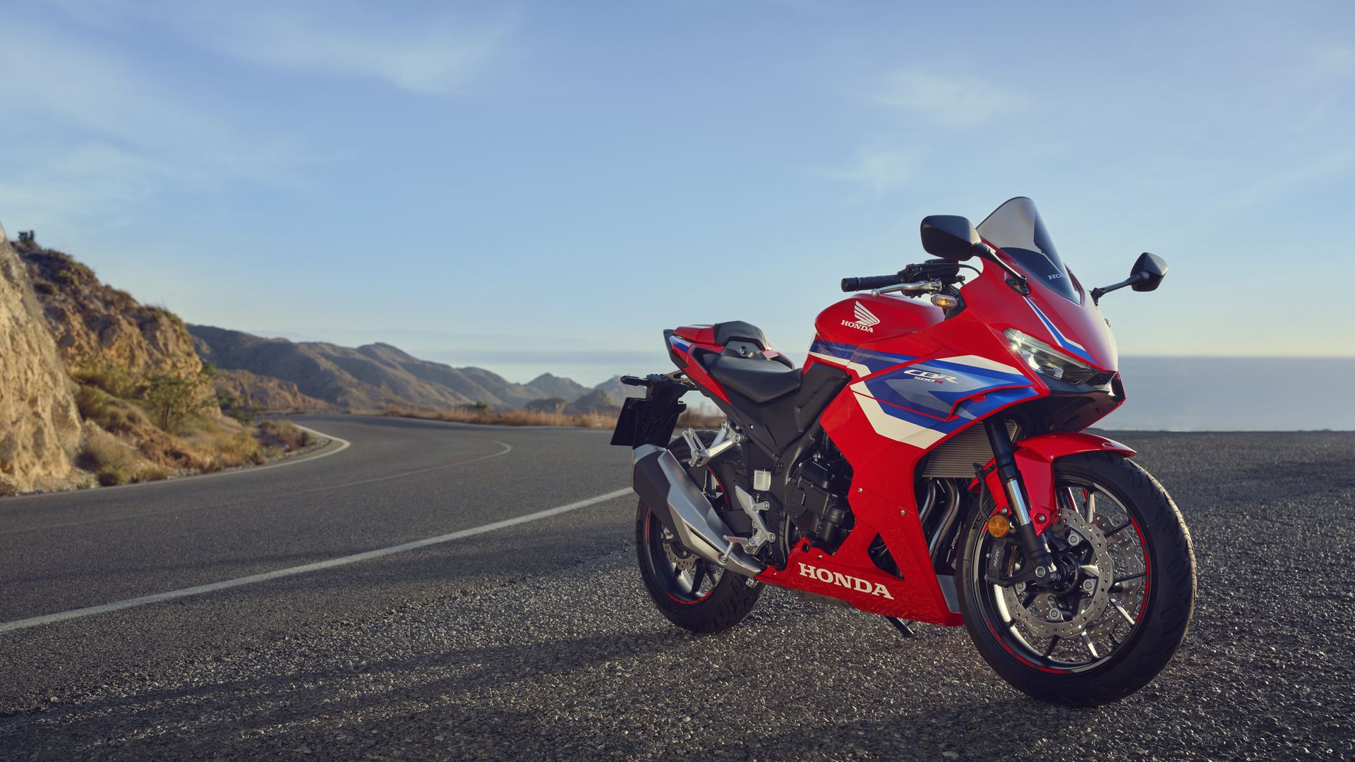 A red Honda CBR500R sportbike parked on a coastal road with mountains and ocean in the background. The motorcycle is facing right, showcasing its sleek design and aerodynamic fairing.