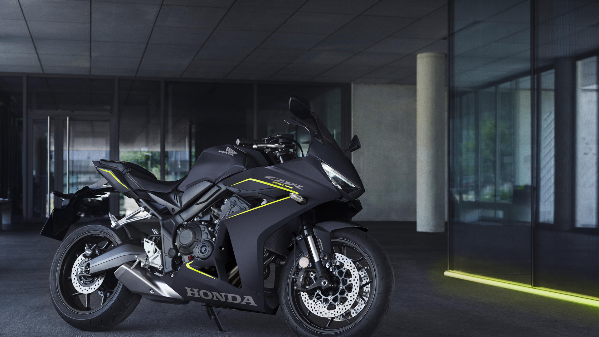 A sleek, dark gray Honda CBR650R sport motorcycle parked indoors in a dimly lit area with glass walls and a concrete ceiling. The bike features yellow accent lines on its fairing and has a sporty, aerodynamic design.