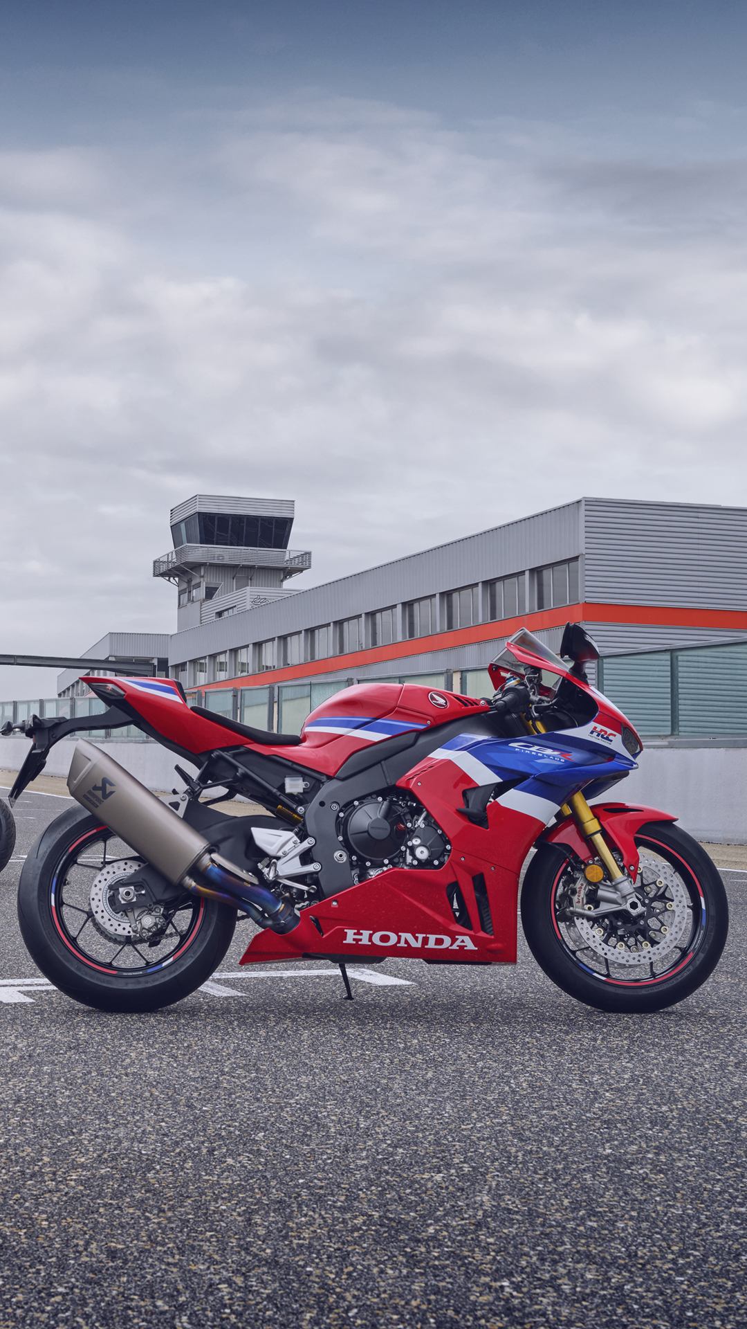 A red, white, and blue Honda CBR1000RR-R Fireblade sport motorcycle parked on a tarmac in front of a gray building with an air traffic control tower, under an overcast sky.