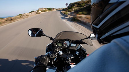 Pohľad cez rameno jazdca na prístroje motocykla, riadidlá a cestu pred motocyklom (exteriér).