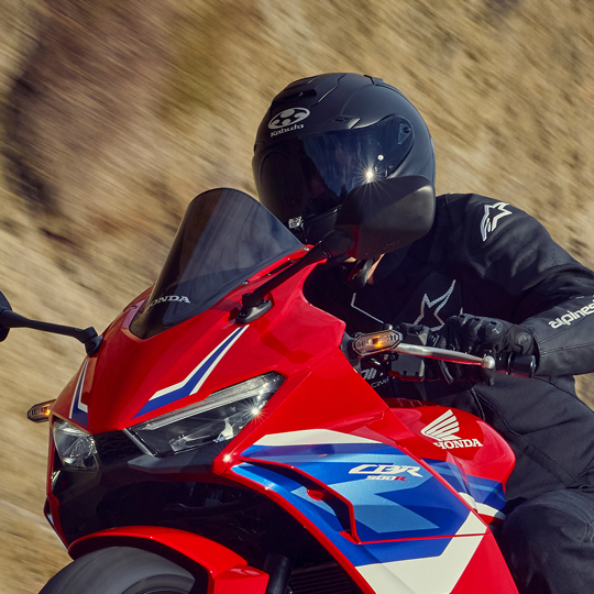 A rider in black gear and helmet on a red, white, and blue Honda CBR motorcycle, leaning into a turn on a blurred background.