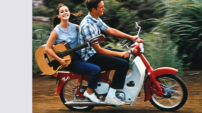 Honda cub with rider, and pillion carrying a guitar. Dirt road location. Right-facing.